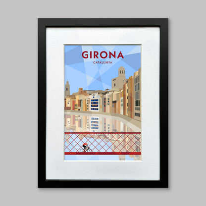 Print of a cyclist crossing the red iron bridge in Girona, Spain over the Onyar River, with the colourful houses of the old city reflected in the water below. Presented in a black frame with a white mount.