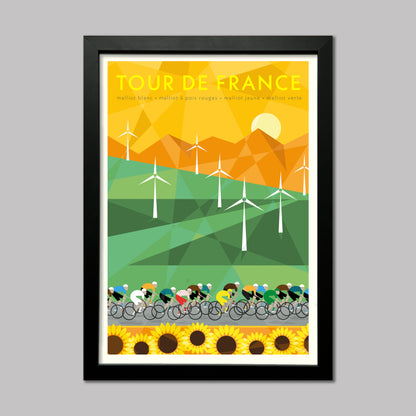 Cycling print of the Tour de France peloton racing past a field of windmills and sunflowers with mountains in the background. The yellow jersey can be seen in the group. Presented in a black frame.